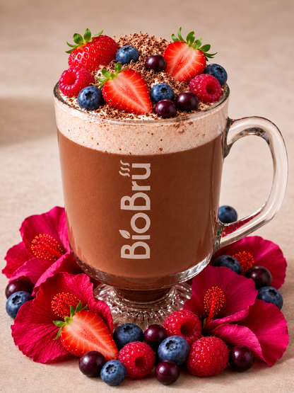 A clear glass mug filled with a pink beverage BioBru bloom, topped with whipped cream, chocolate powder, and fresh berries, arranged on a white surface with flowers and more berries around the base. The mug shows the BioBrú logo.
