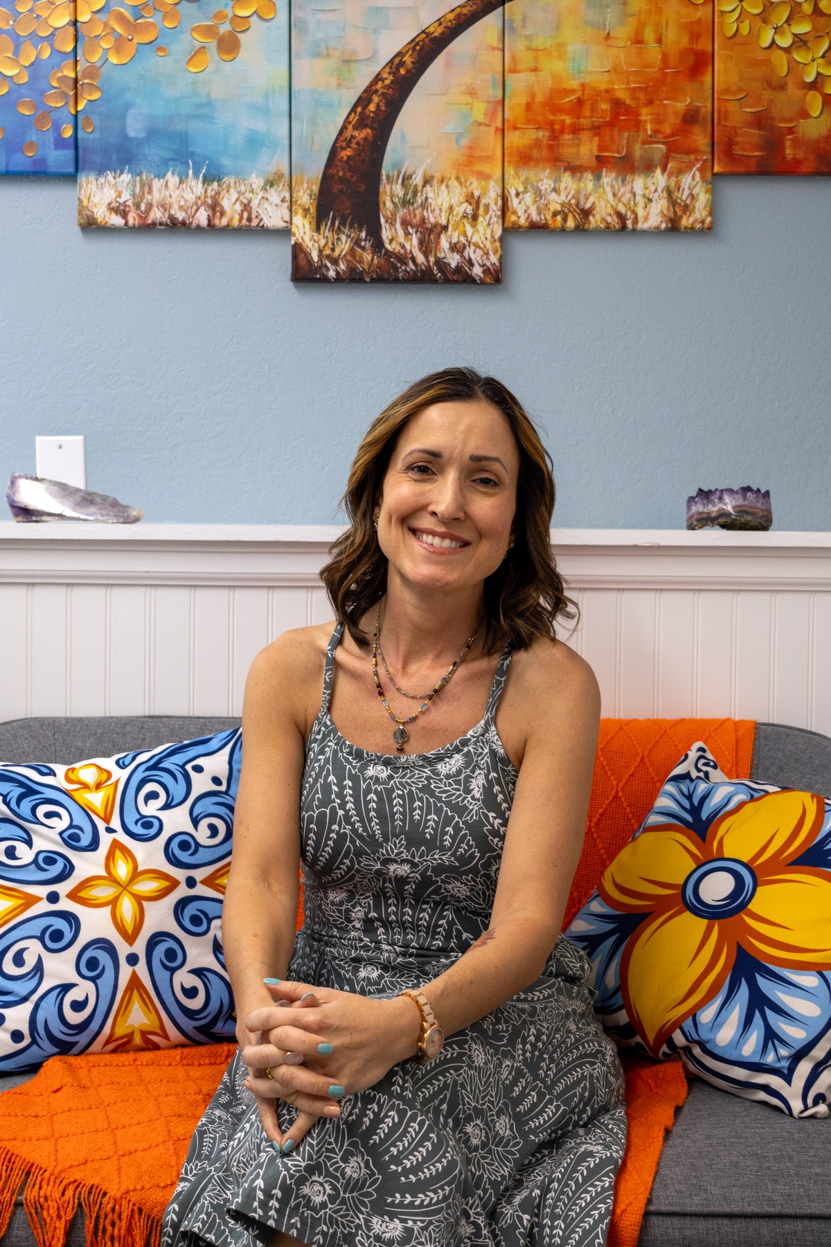 Founder  sitting on a colorful couch with floral cushions, smiling at the camera.